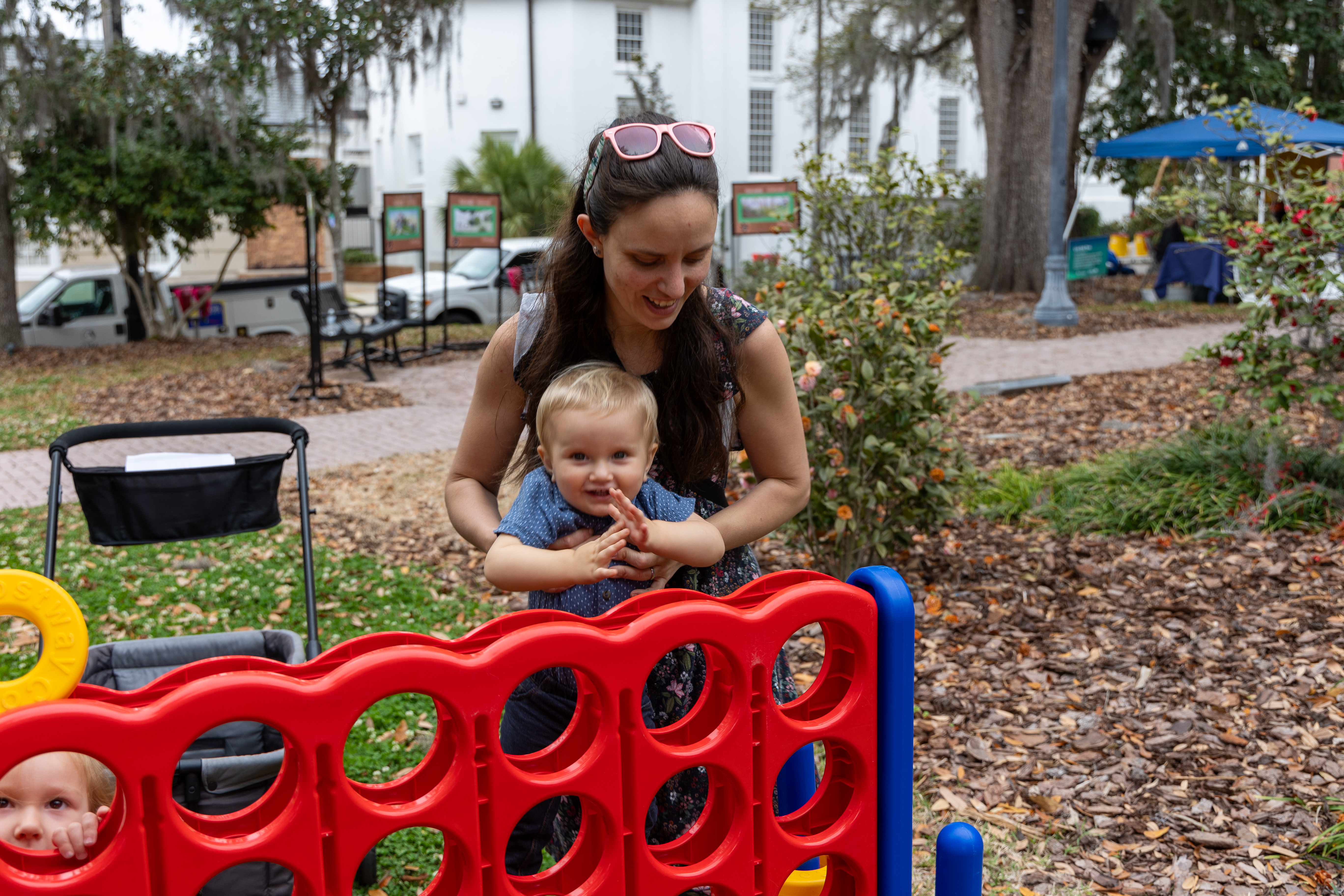 A smiling woman stands behind a happy toddler at a Connect Four game in a park.